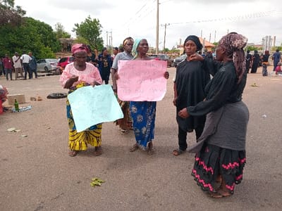 A group of five women stand in a paved, open area, a street, in front of a crowd of people. Four of the women hold signs. The woman on the left holds a white sign with black writing that is illegible. The woman in the center holds a light pink sign with black writing that reads: "We are taking our story as we have a hard time get paid." The two women to the right are not holding signs. The woman in black is gesturing towards the woman standing behind her with a black headscarf, possibly speaking to her. Two more women are visible in the background, walking away from the group. The scene is taking place in front of a building with a large, brown, rectangular facade, and a green tree is visible in the top left corner of the photo. The area is moderately crowded, with cars, people, and a few stray tires on the ground. The scene is located in Ungwan Tanko, Nigeria, at latitude: 10.422 and longitude: 7.49.