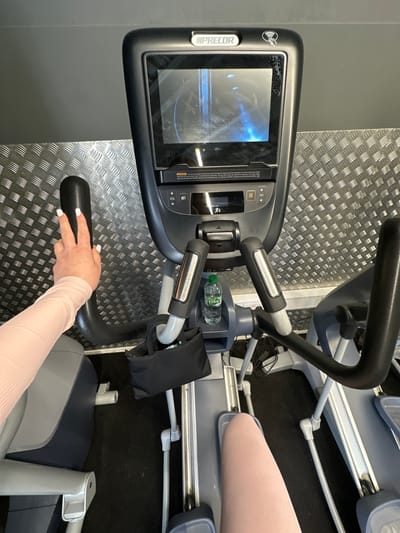 The image shows a close-up view of the console and handlebars of a Precor elliptical machine in a gym. A woman's hand with light pink nail polish is resting on the left handlebar. Her arm is covered by a light pink long-sleeved top. The console displays a blue-toned graphical interface, and a small bottle of water is visible in a holder below the console. The gym environment includes a textured metal wall in the background and black flooring. No specific location in Carlow, Ireland can be determined from the image.
