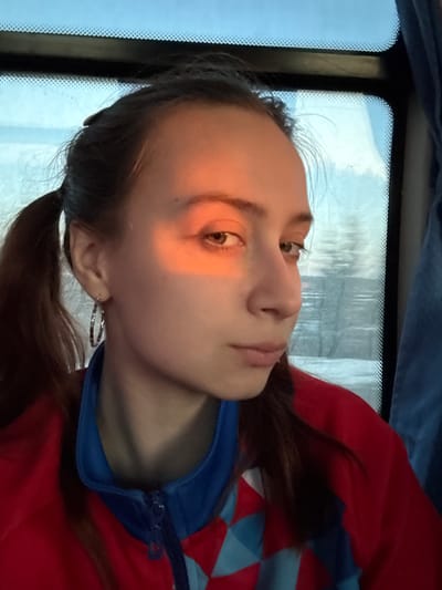 A young woman with light skin and brown hair in pigtails sits by a window with a blurry outdoor scene visible. She wears a red jacket with a blue collar. A bar of sunlight illuminates one side of her face. The image appears to be taken from a low angle, looking up at the subject. Location cannot be identified.