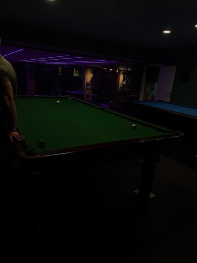 The image captures an indoor scene within a recreational establishment, identified as being in Majdal Shams, Israel. The foreground features the left side of a person, observed from the upper arm to the mid-thigh, wearing a dark olive green short-sleeved shirt. Their right forearm and hand, adorned with a dark bracelet, rest on the dark wooden rail of a billiard table. This table has a green felt surface, on which a black 8-ball is positioned near the foreground left pocket, and a solid white cue ball is located further up the table, closer to the center. In the mid-ground, to the right, a second billiard table with a blue felt surface is partially visible. The background is dimly lit, showcasing sections of an exposed brick wall, horizontal purple LED strip lighting on what appears to be a reflective or mirrored surface, and an open white door with various posters affixed to the adjacent wall. Overhead, multiple circular spotlights are visible, contributing to the establishment's illumination.