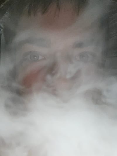 The image shows a close-up, out-of-focus view of a person's face partially obscured by white smoke or vapor. The person appears to be looking directly at the camera. The lighting is dim, and the background is dark and indistinct. There is a reddish-pink area on the left cheek, which could be a blush or a skin discoloration. The overall impression is of a person engaged in an activity that produces a significant amount of smoke, such as vaping or smoking. The image does not provide any information about the location within Jihlava, Czechia.
