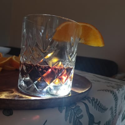 An overhead shot shows a crystal glass filled with a dark red liquid and ice cubes. Two orange slices are perched on the rim of the glass. The glass sits on a wooden tray, which is placed on a tablecloth with a green floral pattern. The scene is lit by natural light, casting shadows and highlights. The background is blurred and appears to be indoors. The image was taken in Limerick, Ireland, specifically in a home setting within the city.