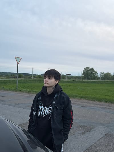 A medium shot depicts a person with short dark hair wearing a black hoodie with a white graphic design and a denim jacket, standing next to a car. They are positioned on a paved road adjacent to a grassy field. In the background, utility poles are visible along with dense foliage. A road sign is also present. The sky is overcast. Based on available data, the location is Azyakul', Russia.
