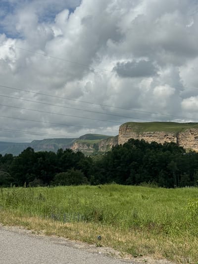 This is a landscape photograph taken in the vicinity of Кубина, Russia. The image depicts a vast, open field of tall green grass in the foreground, with a paved road running along the left edge. Behind the field is a dense line of dark green trees, forming a natural barrier. Beyond the trees, a dramatic series of layered rock formations and hills rise, their sides carved and striated, with a green, grassy plateau capping the uppermost layer. The sky above is filled with dramatic, heavy grey and white clouds, suggesting a dynamic weather system. Multiple power lines are strung across the sky, running horizontally through the frame at varying heights, supported by distant poles that are visible against the landscape. The overall impression is one of rugged natural beauty under a brooding sky.