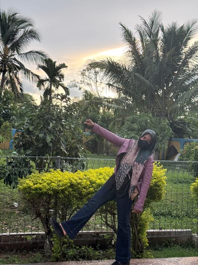 A young woman wearing a pink jacket, a dark hijab, and dark flared jeans stands in a park-like setting with palm trees and lush greenery. She is wearing a face mask and a patterned scarf. Her left leg is extended, and her left arm is raised as if in a playful pose. In the background, a low fence with green mesh encloses the area, and beyond that, a colorful mural with blue and yellow segments is visible. The sky is overcast with hints of sunlight breaking through. The ground is paved with patterned tiles, and there is a small, brightly lit green bush in the foreground. The scene appears to be in Lampeuneurut Blang, Indonesia, given the tropical vegetation and architectural style.