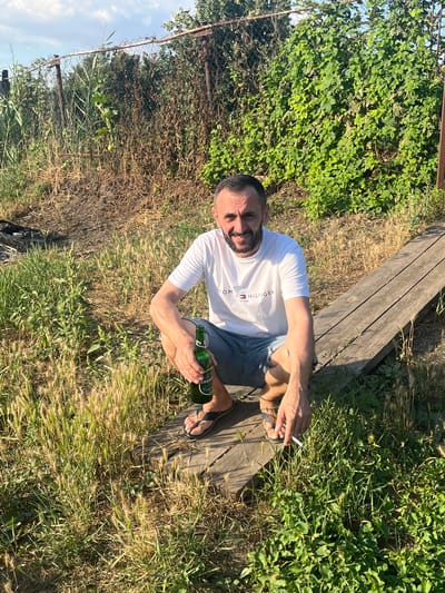 A man with dark hair and a beard is crouching on a wooden plank structure outdoors. He is wearing a white Tommy Hilfiger t-shirt and denim shorts. In his right hand, he holds a green bottle of beer, and in his left hand, he holds a lit cigarette. The background consists of a wire fence, overgrown vegetation, and a clear sky with some clouds. The ground is covered in grass and dry vegetation. The scene appears to be in a rural or semi-rural setting, possibly a garden or backyard. The specific location in the city of Яново-Грушевский, Russia, cannot be determined from the image alone.