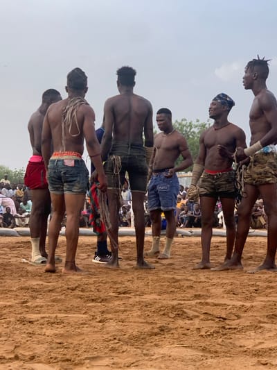 The image shows a group of six men standing on a sandy wrestling pit in Sokoto, Nigeria. They are all shirtless, wearing shorts and some have traditional head coverings. They are facing each other, but are not actively engaged in wrestling. The scene is likely a pre-match preparation or a break during a wrestling tournament. Several people are seated in the background as spectators, watching the men. The sky is overcast, but it is daylight. The coordinates provided place the scene in Sokoto, a city in northwestern Nigeria. The environment is a sandy arena, likely an outdoor wrestling ground.