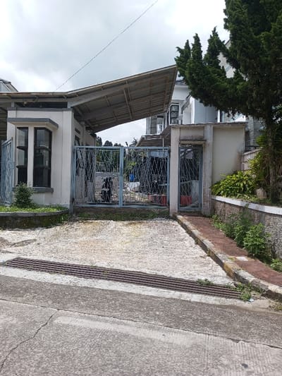 An external view of a residential property entrance in Viengthong, Laos, is depicted under an overcast sky. The foreground shows a grey concrete road with a dark metal drain grate. Beyond the grate, a light-colored concrete driveway leads to a partially open double-leaf metal gate with a decorative diagonal grid pattern. To the left of the main gate stands a small, white-walled building with dark-framed windows and a projecting, sloping metal roof supported by grey beams. To the right of the main gate, a narrower pedestrian gate, also with a grid pattern, is visible, leading to a raised, reddish-brown paved walkway bordered by a low concrete wall and overgrown with vegetation. Inside the property, a black scooter or motorcycle is parked behind the main gate, and various indistinct objects, including what appears to be a blue tarp and red containers, are visible further in. Portions of a multi-story white building with dark-framed windows are visible within the property, behind the gates and adjacent structures. A tall, dark green coniferous tree stands prominently on the right side of the image, within the property. The distant background reveals faint outlines of green foliage or hills beneath the cloudy sky. No individuals are present in the scene.