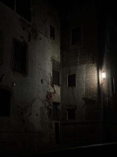 The image depicts a narrow, dark alleyway at night, likely in the historic center of Lagnasco, Italy. Two old, weathered buildings face each other, their brickwork exposed in places where the plaster has crumbled. The building on the left shows signs of disrepair with peeling plaster and visible brick. It features several barred windows, suggesting it may have been a prison or an old fortification. The building on the right is constructed of brick and has a single, lit lantern mounted on its facade, casting a warm glow on the brickwork and the dark alley. The windows on this building are dark, indicating they are either unlit or have shutters. The overall atmosphere is moody and slightly unsettling, with deep shadows and minimal light. There are no people or animals visible in the scene.