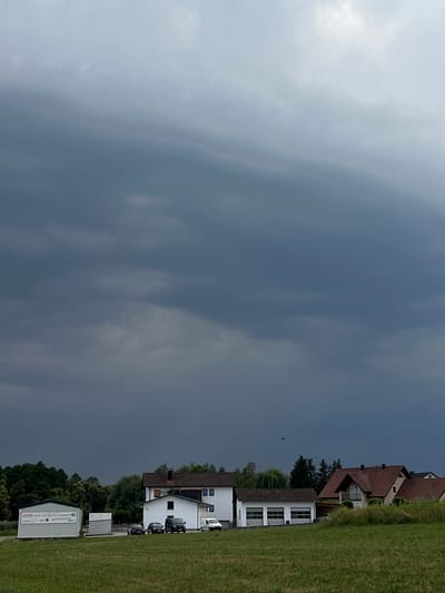 The image depicts a scene in Rudelzhausen, Germany, under a heavily clouded, dark sky. In the foreground, a green grassy field stretches across the bottom of the frame. Behind the field, a line of buildings is visible. To the left, a long, white, single-story building with signage is present, featuring what appears to be a garage door. Adjacent to it is another, smaller white structure. To the right of these, a white two-story house with windows and a dark-colored roof stands. Further to the right, a white garage structure with two large doors is situated. Beyond these buildings, several residential houses with reddish-brown tile roofs are visible. A line of trees forms the background, with some taller evergreen trees interspersed among them. A single dark object, possibly an airplane or bird, is discernible in the sky above the buildings. The overall atmosphere suggests an impending storm or overcast weather.