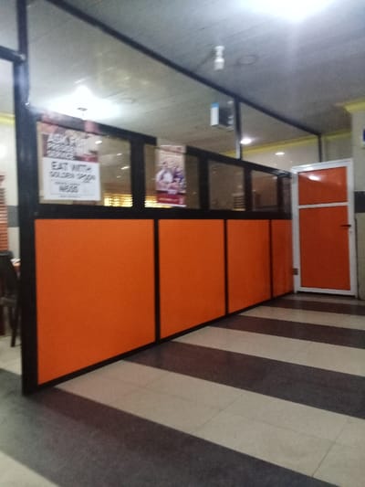 The image shows a view of an interior space in a restaurant located at the coordinates 5.503 latitude and 5.799 longitude in Ovwian, Nigeria. The space appears to be a waiting area or reception area, with a black-framed glass partition separating it from another section of the restaurant. The partition has orange-colored panels on the bottom. In the background is a closed door, also orange, framed in silver. The floor is tiled with alternating black and white stripes. On the wall of the partition, a poster promoting “Golden Spoon” with the slogan “Eat with the Golden Spoon” can be seen. There are no people or objects in the space except for the partition and door.  The lighting is bright.  The image is taken from a low angle, looking toward the partition.