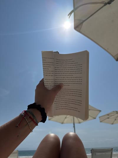 A first-person point of view image captures a leisure scene on a sunny day in La Cruz de Huanacaxtle, Mexico. The subject's tanned thighs and lower legs are visible in the foreground, indicating direct sunlight. A left hand, adorned with multiple multi-colored string bracelets and a black scrunchie, holds open a paperback book titled "Things We Never Got Over" by Lucy Score. The book's pages display English text, with page number "3" visible on the right. The background features a clear, bright blue sky with a strong sun directly overhead, causing a lens flare. Two light-colored beach umbrellas are partially visible, providing shade in the middle ground, with the ocean faintly discernible in the far left background. The overall environment suggests a beach or resort setting.