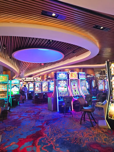 The image shows an interior view of a casino on the Hollywood Seminole Reservation in the United States of America. The scene is dominated by rows of slot machines with illuminated displays and neon accents. A patterned carpet in shades of blue, orange, and red covers the floor. Along the left side is a curved architectural feature with wooden slats and a circular illuminated element. Several people are visible, either standing or sitting at the slot machines. The overall lighting is vibrant and colorful, creating a lively atmosphere.