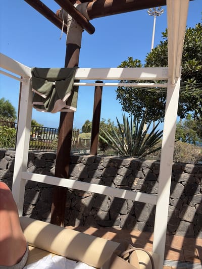 The media file displays an outdoor scene, likely a patio or resort area, characterized by a wooden and white-painted structure that frames the view. The scene is bathed in bright sunlight, suggesting a daytime setting. A large, spiky agave plant is prominent in the middle ground, set against a background of dark stone walling and lush greenery. Beyond the immediate surroundings, a glimpse of the ocean or a large body of water is visible, hinting at a coastal location.

In the foreground, parts of a person's leg and a beige handbag are visible, indicating someone is present, possibly relaxing. A green towel with cream trim is draped over one of the wooden support beams of the structure. Shadows cast by the structure and surrounding foliage create a dynamic play of light and dark across the stone wall and the ground. The overall mood appears tranquil and summery, consistent with a holiday or leisure environment. The location is identified as Antigua, Spain. There is no visible text in the media file.