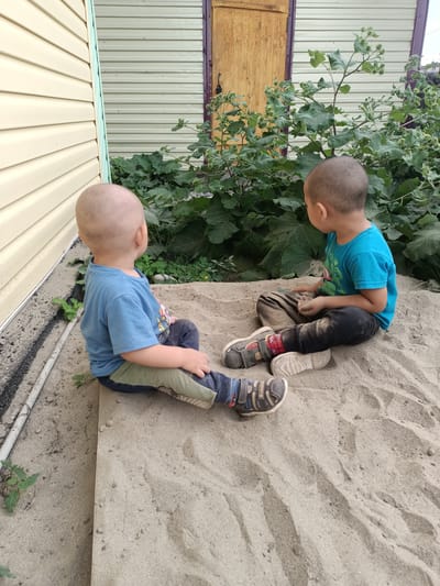 Two young boys are sitting in a sandbox in a yard. The boy on the left is wearing a blue t-shirt and green and blue patterned pants. The boy on the right is wearing a blue t-shirt and dark pants. Both boys are barefoot except for the boy on the right, who is wearing shoes. The sandbox is filled with sand, and there are some plants growing around it. In the background, there is a building with a wooden door and siding. The scene appears to be outdoors on a sunny day. There is no visible text in the image.
