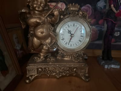 In the city of Yazykova Pyatina, Russia, a close-up shot focuses on an ornate, gold-colored clock with a cherubic figure holding a violin as part of its design. The clock face features Roman numerals and black hands, with the time appearing to be approximately 9:33. The clock is set upon an intricately carved golden base, adorned with floral motifs. To the left of the clock, a framed picture of a person wearing a white headdress is partially visible. To the right, the lower portion of a doll or mannequin dressed in dark clothing with purple accents and black boots is in the frame. The background consists of a dark, blurred tapestry-like pattern with hints of floral designs in shades of pink and red. The scene is lit, illuminating the golden hues of the clock and its stand.