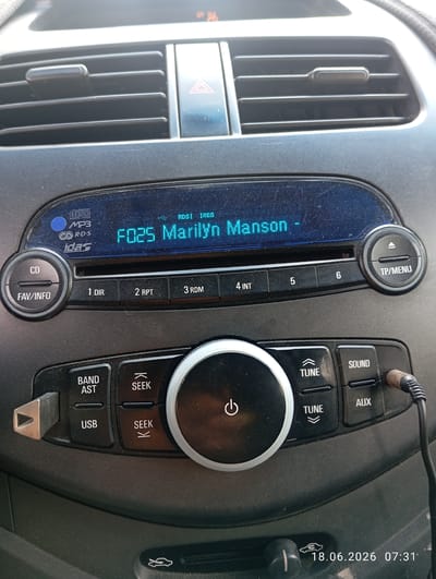 An interior car dashboard is shown, with a close-up view of the car's audio system. The system displays "F025 Marilyn Manson" on its screen, indicating a song is playing. Buttons for "CD", "FAV/INFO", "1 DIR", "2 RPT", "3 RDM", "4 INT", "5", "6", "TP/MENU", "BAND AST", "USB", "SEEK", "TUNE", "SOUND", and "AUX" are visible. A power button with a symbol of a circle and a vertical line is prominent on a central dial. An audio jack is plugged into the "AUX" port. The date and time "18.06.2026 07:31" are superimposed on the image. The context of Ohangaron, Uzbekistan, is not visually evident from the image itself, but is provided as external information.