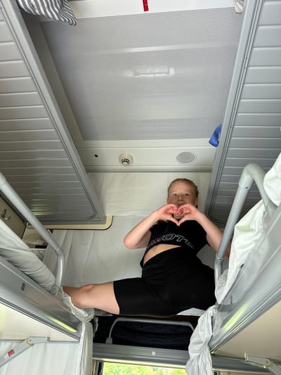 The image, rotated clockwise, captures a young person posing inside what appears to be a train sleeper car or a similar compact bunk bed accommodation. The person, with reddish hair and dressed in a black t-shirt and shorts, is seen from the waist up, making a heart gesture with their hands towards the camera. Their t-shirt displays partially visible text that appears to read "VOTE".
The setting features tightly packed bunk beds with metal frames and white curtains or privacy screens. One bunk bed is prominent in the foreground and lower left, with another partially visible above and to the right. The walls are light-colored, with a large section on the left featuring a perforated white panel. A small, circular vent or speaker is visible on the wall behind the person. On the left edge, a red and white warning label in Cyrillic text is partially visible, stating "ОПАСНО" (DANGER) and "ЭТО ОПАСНО" (THIS IS DANGEROUS).
Through a window on the right side, lush green foliage is visible, suggesting a daytime scene in a natural environment. The overall impression is one of travel, likely within a train compartment in Васильево, Russia, given the provided location context and the design of the bunks. The person's pose indicates a playful or cheerful mood.