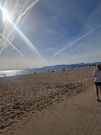 The image shows a beach scene in Les Meravelles, Spain. The perspective is tilted. The sky is blue with several contrails from airplanes. Below the sky, the ocean shimmers with sunlight. The beach stretches to the right, with people scattered along the shore. A boardwalk runs parallel to the beach, and a person is walking along it. In the distance, a mountain range is visible. The sand shows footprints and patterns.