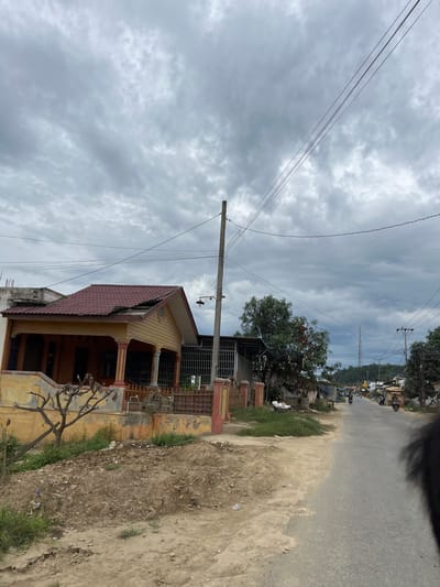 The image captures a street scene in Rantau Pauh, Indonesia, under a heavily overcast sky. A modest, yellow and red-tiled house with a covered porch is visible on the left side of the frame. The house appears to be in a state of disrepair, with a broken section of the roof. Adjacent to the house, a bare tree stands in a patch of overgrown grass and dirt. A paved road extends into the distance, with several motorbikes and pedestrians traveling along it. The roadside is lined with various structures, including small shops and other houses, and interspersed with trees and utility poles supporting overhead wires. A tall transmission tower is visible in the background, silhouetted against the grey sky. The ground along the road is a mix of dirt and gravel.