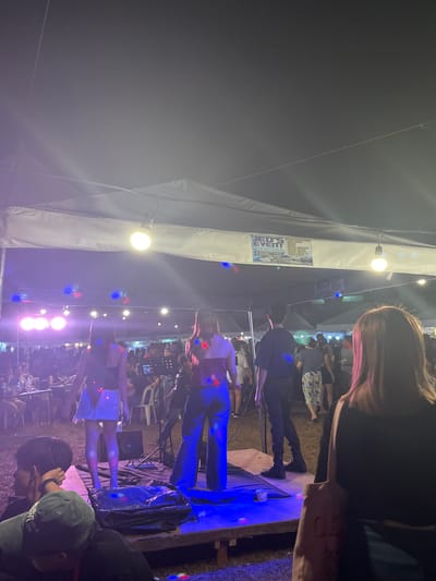 The image depicts an outdoor evening event in Koronadal City, Philippines, possibly a performance or a gathering held under a large white tent. The lighting is predominantly purple and blue from stage lights and possibly disco balls, creating a vibrant atmosphere. In the foreground, three people are visible from behind, with the closest being a woman with brown hair looking towards a stage.
On the stage, three individuals are performing or preparing to perform. A woman in the center is wearing a black top and wide-leg blue pants, facing away from the camera. To her left is another woman in a black top and a short, light-colored skirt. To the right of the woman in blue pants is a man in a black shirt and black pants, facing the audience.
Behind the performers, the crowd is visible, with many people seated at tables or standing. There are additional tents and lighting structures in the background. A banner reading "JED'S EVENT RENTAL" with contact information is visible on the tent above the stage, suggesting this is a rented event space. The ground appears to be grass or dirt, typical of an outdoor venue. The overall scene conveys a lively and festive ambiance.