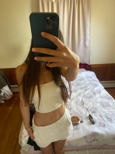 A young woman is taking a mirror selfie in a bedroom. She is wearing a white tube top and a white lace skirt. She has long brown hair and a belly button piercing. The room has wood floors, a bed with floral bedding, and a radiator. There are curtains on the wall and a dresser with clothing on it. The image was taken in Oak Bluffs, United States of America.