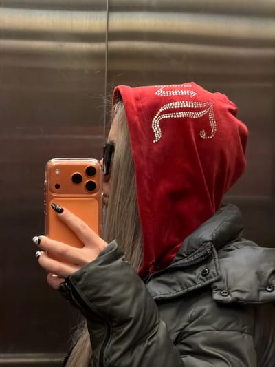A person wearing a red velvet hooded scarf with a large, jeweled "JC" logo is holding an orange-cased smartphone. The person is also wearing a gray puffer jacket. The background appears to be a metallic surface. The exact location in Bağcılar, Turkey is not discernible from the image.