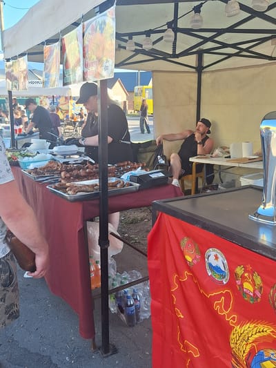 At an outdoor market in Цімкавіцкі сельскі Савет, Belarus, a food stall displays trays of grilled meats and sausages. Two vendors, a woman in a black cap and apron and a man wearing a black tank top and cap, are present. The woman is behind the counter, seemingly preparing food, while the man sits in a chair, looking thoughtfully to the side. Various beverages are visible in plastic bags and on shelves. A red banner with Soviet-era emblems and a map of the USSR hangs from the front of the stall. The background shows other market stalls, people, and a yellow bus. The overall scene depicts a bustling outdoor food vendor operation.