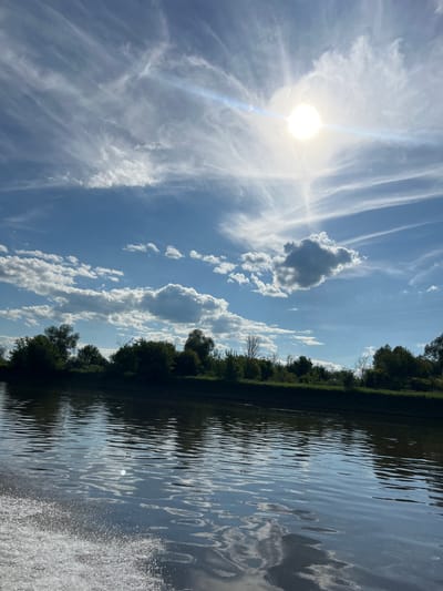 The image depicts a tranquil scene of a body of water, likely a river or lake, with a lush green tree line bordering its edge. The water's surface is rippled, reflecting the bright sky and the surrounding foliage. The sky is a vibrant blue, dotted with a mix of wispy cirrus clouds and larger cumulus formations. The sun is visible, casting a bright, diffused light, suggesting it is daytime, likely mid-morning or afternoon given the sun's position and intensity.
The tree line appears dense and healthy, composed of various deciduous trees, indicating a natural, undeveloped setting. The presence of the water and trees together suggests a riparian environment. The reflections in the water are detailed, showing distorted images of the clouds and the trees, adding depth to the composition.
There are no people or man-made objects prominently featured in the image, emphasizing the natural beauty of the landscape. The overall mood is peaceful and serene, evoking a sense of calm and connection with nature. The scene is likely captured in a rural or natural park setting. The image is oriented vertically.
Given the location context of Ust'-Liuga, Russia, this scene could be representative of the natural landscapes found in that region during the warmer months, possibly during summer or late spring. The weather appears to be fair and sunny, with some cloud cover. The time of day is clearly during daylight hours. There is no visible text in the image.