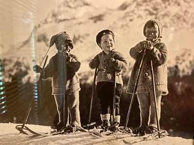 A black and white photograph captures three young children dressed in winter ski attire, standing on a snowy slope with skis and poles. The children, appearing to be siblings or close friends, are positioned from left to right. The child on the left wears a knitted hat with a pointed tip and a textured sweater. The middle child sports a brimmed hat and a sweater with a belt. The child on the right is also wearing a knitted hat and a patterned sweater. All three are holding ski poles.
The background suggests a mountainous or hilly terrain covered in snow, with some trees or rocky formations visible. The lighting indicates it might be daytime, possibly a sunny winter day, given the bright highlights on the snow and the subjects. The children's expressions are cheerful and engaged, with the middle child looking up and smiling, and the other two also appearing happy. The scene conveys a sense of playful winter activity. The location context provided is Ленинское, Russia, suggesting this is a winter scene from that region. There is no visible text in the image.
