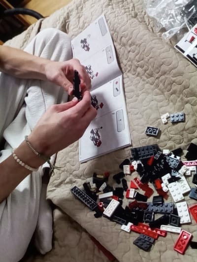 The image depicts an indoor scene in the city of Brzezina, Poland, showing a person engaged in assembling a model with building bricks. The individual, visible from the mid-torso downwards, is wearing light-colored, loose-fitting trousers. Their left wrist is adorned with two bracelets: one composed of small, light-colored beads and another thinner, cord-like bracelet. The person's hands are centered in the frame, holding and connecting black interlocking plastic bricks, forming a partially assembled structure. An open instruction booklet, displaying sequential assembly diagrams, lies on a quilted, light-brown textured surface to the left of their hands. To the right, a scattered pile of various black, white, and red interlocking plastic bricks is visible, along with a translucent plastic bag partially obscured in the upper right, likely containing additional parts. The activity takes place on a quilted surface, possibly a bedspread or blanket, with a ripple-like textured pattern. The background on the upper left is dark and indistinct.