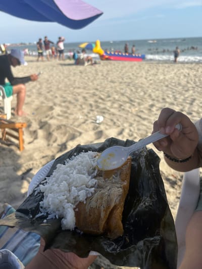 The image displays a bustling beach scene in General Villamil Playas, Ecuador, under a clear blue sky. In the immediate foreground, a hand holds a white plastic plate on which a portion of food wrapped in dark green leaves is served, topped with white cooked rice. A second hand holds a white plastic spoon with yellowish residue, positioned over the dish. To the left, a man wearing a white cap, a dark long-sleeved shirt, and light-colored shorts sits on a white plastic chair beneath a partially visible purple and blue beach umbrella, intently viewing a smartphone. A small wooden stool is next to his feet on the sand. The midground and background consist of light-colored sand and the ocean. Numerous people are scattered across the beach and in the water. A large, inflatable red and blue banana boat with yellow accents is visible in the ocean, alongside other smaller vessels and swimmers.