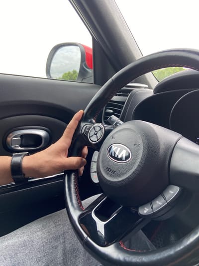 In Pervomaiskoe, Russia, a person's hand with a black wristwatch is seen resting on the steering wheel of a car. The steering wheel has red stitching and the Kia logo is visible in the center. A portion of the car's interior, including the door handle and dashboard, is also visible, along with a side mirror showing a cloudy sky and trees. The person appears to be driving, or about to drive, the vehicle.