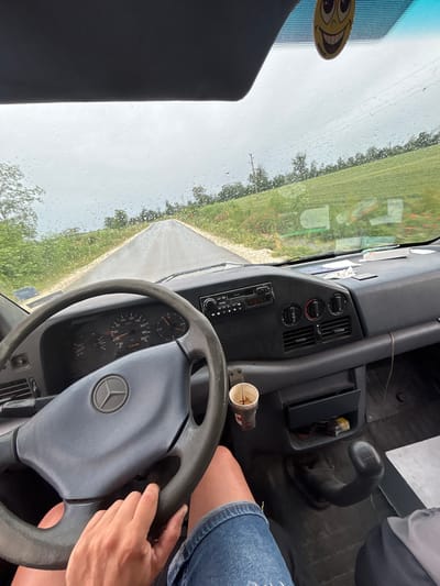 The image provides a first-person view from the interior of a vehicle, looking through a windshield dotted with raindrops. A person's hands are gripping the steering wheel, and their bare leg, wearing denim shorts, is visible to the right. The steering wheel features a Mercedes-Benz logo, and the dashboard is primarily dark gray and black.

The instrument cluster behind the steering wheel shows a speedometer reading up to 200 km/h, a tachometer, and an odometer displaying 575,389. Below the central dashboard, a radio with "PHILIPS" and "ENERGY" visible on its face is present, flanked by air vents and climate control knobs. To the right, a manual gear stick is prominent, and near it, a used paper cup sits in a holder. A yellow, oval-shaped sticker with a wide-eyed, smiling face is attached to the upper left corner of the windshield.

Through the rain-streaked windshield, a narrow, unpaved or dirt road is visible, stretching into the distance. Lush green fields and scattered trees line both sides of the road, suggesting a rural environment. The sky appears overcast and grey, consistent with the rain. The overall setting implies a drive through the countryside in Dobrotitsa, Bulgaria, likely during daytime and in wet weather conditions.