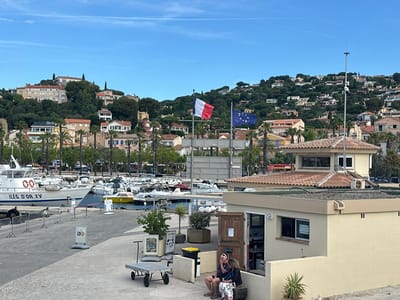 The image captures a view of the port of Le Lavandou, France, with the town nestled on a hillside in the background. In the foreground, a beige building with a tiled roof houses what appears to be a service or information point. Two individuals are seated on a low wall outside this building, engaged in conversation. A hand trolley is parked nearby, along with various bins and signage.

The middle ground of the scene is dominated by a marina filled with numerous boats of varying sizes, moored in the clear blue water. The largest boat in view bears the inscription "ILES D'OR XV". A yellow boat is also visible. Palm trees line the waterfront, adding a tropical feel to the Mediterranean setting.

In the background, the town of Le Lavandou sprawls across a green, tree-covered hillside, with numerous houses and buildings cascading down towards the sea. The French and European Union flags are prominently displayed on flagpoles, fluttering in the breeze against a clear blue sky. The overall impression is one of a bustling seaside town on a pleasant, sunny day.