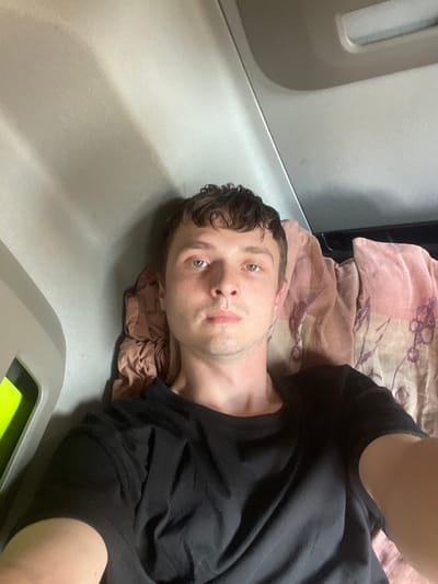 A man lies on his back on a flat surface, likely a bed or cot, in what appears to be a confined space. He is wearing a black t-shirt. The surface he is lying on is covered with a patterned fabric, possibly a blanket or sheet, with colors of pink and brown. Beside him, the wall of the space is visible, made of a light gray material. To the left of the man, a portion of a white vertical structure with a curved indentation is visible, suggesting it might be part of a wall or a piece of furniture. In the lower right corner of the image, a gray object with a bright green section is partially visible, the purpose of which is unclear from the image. The man's eyes are open and he is looking directly at the camera, with a neutral expression. The lighting suggests it could be daytime or a well-lit indoor environment. There is no visible text in the image.