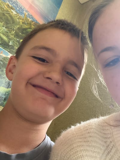This is a close-up selfie taken by a young boy, possibly with an adult in the background. The boy is smiling broadly, with his eyes looking directly at the camera. His hair is short and brown. He is wearing a grey t-shirt. The background is blurred, but appears to be a living room with a green textured wall and a large picture or tapestry with a landscape scene featuring a body of water and foliage. Part of a cream-colored fuzzy sweater is visible in the upper right corner, suggesting the presence of another person, likely an adult. The lighting suggests it is daytime, possibly indoors near a window. The overall emotion conveyed is one of happiness and playfulness. There is no visible text in the image. The image was taken in Krasnovka, Russia.