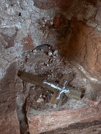 The image displays a close-up view of a crucifix nestled within a rugged, cave-like or grotto-like natural rock formation. The central object is a small crucifix, composed of a weathered wooden cross with a metallic, silver-colored corpus of Jesus Christ affixed to it. Jesus is depicted with outstretched arms, a halo around his head, and a draped loincloth.
The setting is characterized by uneven, textured rock surfaces predominantly in shades of reddish-brown, dark brown, and grayish-white. The rocks exhibit numerous cracks, crevices, and granular patches, suggesting a natural and possibly ancient environment. Loose pebbles and rock debris are visible on ledges and at the base of the crucifix, blending into the earthy surroundings.
No activity is taking place; the scene is a still life, presenting a devotional object in its environment. The lighting is diffused, suggesting an interior space or a sheltered area, rather than direct sunlight. There are no visible signs of specific time of day or weather conditions. The placement of the crucifix evokes a sense of solemnity and reverence, characteristic of a religious shrine or a personal devotional spot. The location context indicates this is in Staroe Rakomo, Russia. No visible text is present in the image.