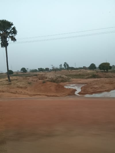 This image captures a rural landscape in Barakin Kuru III, Nigeria, under an overcast sky. The foreground presents a broad, blurred reddish-brown surface, consistent with a dirt road or track viewed from a moving vantage point. The midground features undulating terrain composed of reddish-brown earth, marked by multiple mounds of disturbed soil and patches of dry, light-colored vegetation. A winding, light-colored stream or pooled water body is visible within this disturbed earth. On the far left, a tall, slender palm tree with a dark trunk and green fronds stands prominent. Three parallel power lines traverse the upper portion of the sky horizontally. The background reveals a distant horizon line with a variety of green trees and shrubs, alongside faint structures resembling utility poles. No human subjects or discernible actions are depicted.