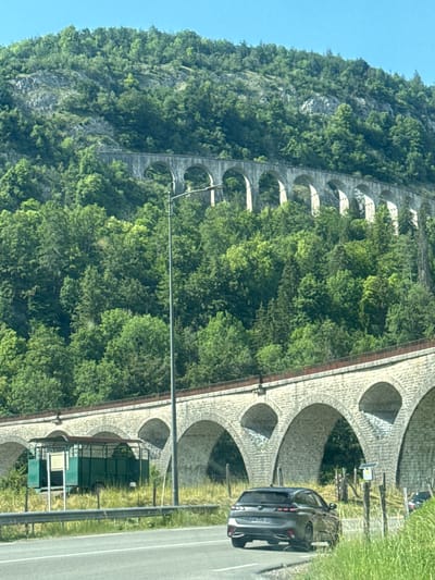 This image, taken in the vicinity of Hauts de Bienne, France, captures a scenic view of a multi-tiered stone viaduct carved into a forested mountainside under a clear blue sky. The foreground features a paved road with a dark grey SUV driving away from the viewer. To the left of the road, a green, open-sided trailer with a canopy is parked on a grassy embankment, adjacent to a metal guardrail. The prominent viaduct, constructed from light-colored stone arches, spans across the frame, with a second, higher viaduct visible further up the slope. Dense green trees cover the mountainside, with some rocky outcrops interspersed. A tall, slender lamp post stands near the lower viaduct on the right side of the road. The overall environment is rural and mountainous, characterized by extensive vegetation and historic architectural structures.