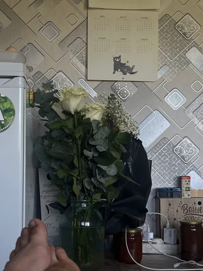 A close-up shot captures a bouquet of white roses and eucalyptus in a glass vase, set against a patterned wallpaper. To the left, a portion of a white refrigerator is visible, featuring a green circular sticker. A calendar hangs on the wall, displaying months and a cartoon cat. In the foreground, a human foot is positioned towards the lower left corner. The background reveals various objects including jars, electrical outlets, and a box with text. The overall setting appears to be an indoor domestic environment.