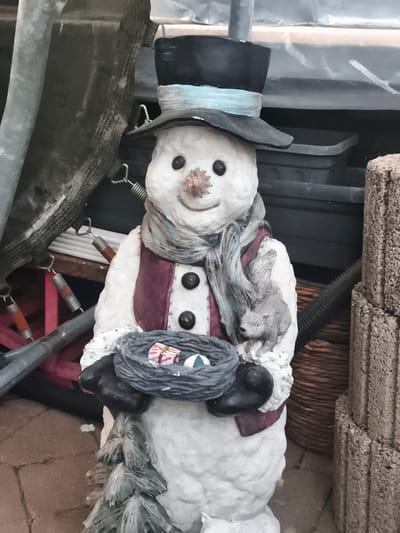 A static, full-body frontal medium shot captures a decorative snowman figurine in an outdoor setting. The snowman, made of a white, textured material, wears a black top hat with a light blue ribbon, a purple vest with three black buttons, and a gray scarf. Its eyes are black dots, and its nose appears to be a small, reddish-brown carrot or berry.

The snowman holds a dark gray, woven nest in its gloved hands. Inside the nest are two small, brightly wrapped gift boxes with red and white stripes, and a green and white striped ball. A small, gray squirrel figurine is perched on the snowman's right arm, its tail wrapped around the arm.

The background is cluttered and appears to be an outdoor storage area or shed. To the left of the snowman, a thick gray pipe or pole runs vertically, partially obscured by a dark, textured material. Several metallic springs with orange ends are visible in the mid-ground. Behind the snowman, gray plastic storage bins are stacked. To the right of the snowman, a rough-textured, beige block structure is visible.

The ground beneath the figurine is paved with reddish-brown brick pavers. The lighting suggests it is daytime, with diffused light. The image's resolution is moderate, with some visible digital noise. The snowman figurine is positioned in the center of the frame, making it the primary subject. No people are present in the image. The scene suggests a decorative display, possibly for a holiday or seasonal event, situated in a somewhat unkempt outdoor environment. The location is identified as Eitorf, Germany.