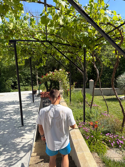 A young man with short hair and sunglasses walks down a set of stone stairs. He is wearing a white t-shirt with a wing design on the back and blue swim trunks. The man is walking away from the camera, under a trellis covered in grapevines. To the right of the stairs is a garden with various flowers and greenery. The background shows trees and a fence. The location is the city of Chavei, Bulgaria.
