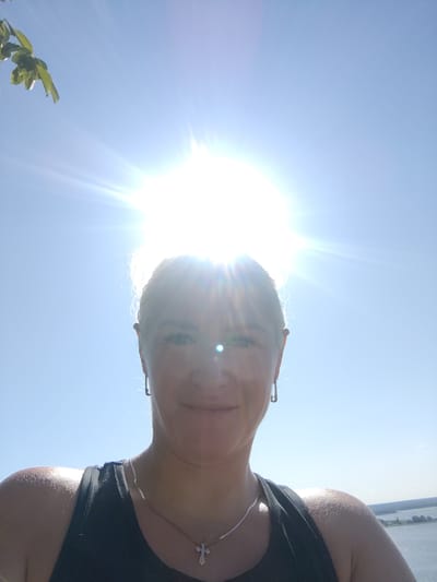 This is a selfie of a woman taken outdoors on a sunny day. She is looking directly at the camera, with the sun positioned behind her head, creating a bright flare. The woman has blonde hair, light-colored eyes, and is wearing a black tank top, hoop earrings, and a silver necklace with a cross pendant. Her right shoulder and arm are visible, and her skin appears to be glowing from the sunlight. In the background, a body of water is visible with a distant shoreline. The sky is clear and blue.