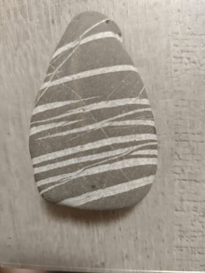 A smooth, oval-shaped gray stone with parallel white stripes lies on a light-colored, textured surface. The surface appears to be wood grain or a similar material, with visible lines and variations in tone. The stone is positioned slightly off-center, with its wider end towards the top of the image and tapering to a point at the bottom. The white stripes on the stone are irregular and vary in thickness, suggesting a natural formation. The lighting is even, casting no distinct shadows, indicating an indoor or overcast environment. There are no people, animals, or specific landmarks visible that would indicate a location in Nor Kharberd, Armenia.