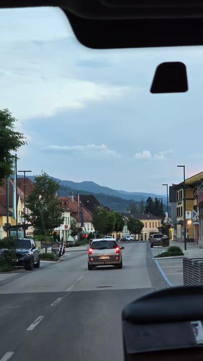 The image shows a view from inside a vehicle looking out onto a street in Kindberg, Austria. The sky is cloudy with a mix of light and dark clouds. On the left side of the street, there are buildings with red tiled roofs and trees. On the right side of the street, there are more buildings, including one with a yellow facade and a sign with a red circle and a white 'H' symbol, indicating a hospital or health facility. A street lamp is visible on the right.

Several vehicles are on the road. In the foreground, the rear of the vehicle the photo is taken from is visible. Further down the street, a silver station wagon with its brake lights on is in the center lane. To the left of the station wagon, a dark colored SUV is parked on the side of the road. Further down the street, another dark colored SUV is parked. A motorcycle is parked near the sidewalk on the left. In the background, rolling hills covered in trees can be seen. The overall atmosphere is calm, likely during the evening or overcast day.