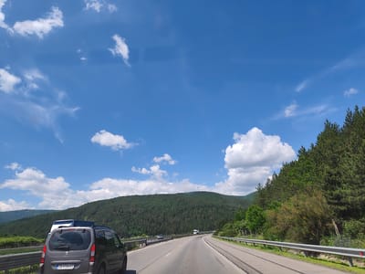 This media file captures a scenic drive through a mountainous landscape in Potop, Bulgaria. The primary subjects are the road, the surrounding nature, and vehicles on the road. The scene depicts a bright, sunny day, suggesting it is daytime.
The foreground features the rear of a grey Peugeot Partner Tepee, partially obscuring a white vehicle ahead of it, indicating a journey in progress. The road ahead is a paved highway, bordered by metal guardrails and a grassy verge. On the right side of the road, a dense forest of evergreen trees climbs the side of a hill. On the left, the landscape is characterized by rolling hills covered in a mix of trees and greenery, with distant mountains visible under a vast blue sky dotted with white, fluffy clouds. A prominent feature on the far hill is a tall communication tower.
The weather appears clear and pleasant, suitable for travel, with ample sunlight casting shadows and highlighting the textures of the landscape. The overall emotion conveyed is one of peaceful travel and appreciation for natural beauty.
Visible text includes "PEUGEOT" and "PARTNER TEPPEE" on the back of the grey car. The license plate reads "CB 4106 KT".