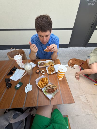 A young man in a blue checkered shirt is seated at a wooden table, eating a hamburger. He is looking down at his food with a slight smile. On the table in front of him are several fast-food items, including another hamburger, french fries, chicken nuggets, waffle fries, and two cups of drinks. A brown paper bag and several napkins are also on the table, along with a pair of black sunglasses and a small green object, possibly a hand sanitizer.
In the background, a person is seated on a bench, their legs crossed, wearing a light-colored skirt and sandals. A small, reddish-brown dog is lying down next to them, tethered by a red leash. The setting appears to be an outdoor seating area of a fast-food restaurant. The floor is tiled, and a portion of a building with textured walls and dark vertical and horizontal lines is visible. The overall impression is a casual meal outdoors.
There is visible text on the building in the upper left corner that reads "SLUZBENI ULAZ", which translates to "OFFICIAL ENTRANCE" in Serbian. This suggests the location is in Serbia, likely Ub Municipality based on the provided context. The lighting suggests it is daytime, with no clear indicators of the weather conditions. The mood seems relaxed and informal.