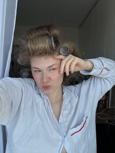 A young woman with blond hair, partially styled with hair rollers, poses for a selfie. She is wearing a light blue and white striped button-down shirt with red trim. Her hand is near her forehead. The background appears to be a room with a window. Cables and other items are visible on a surface behind her. The exact location in Tashevka, Russia, cannot be determined from the image.