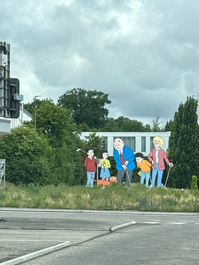 The image, taken in Zurzach, Switzerland, presents a vibrant roadside display featuring several cartoon-style cutouts of people playing golf. The scene is set on a grassy verge adjacent to a paved road, with lush green trees and a modern building in the background.

The cutouts portray what appears to be a family enjoying a golf outing. From left to right (when oriented correctly): an adult woman with blonde hair, wearing a red jacket and blue pants, holds a golf club. Beside her, a young boy with black hair in an orange shirt and blue pants looks on. A central figure is a bald adult man in a blue jacket, white shirt, and red tie, also holding a golf club. Further right, a girl with black hair, dressed in a yellow-green top and a blue-and-white striped skirt, poses with a golf club, accompanied by a small, reddish-brown animal. Another smaller individual in a red-brown top and blue pants holds a club, completing the golfing ensemble. All characters exhibit cheerful, smiling expressions.

The sky is overcast with light grey clouds, indicating a cloudy daytime. Behind the figures, verdant trees and bushes create a natural backdrop, and a contemporary, light-colored building with large windows is partially visible. The right side of the image features a multi-lane paved road with clear white markings. On the far left, parts of a large vehicle, possibly a truck or trailer, and some industrial equipment can be seen. This display appears to be a public art installation or an advertisement, likely related to a golf attraction in the vicinity. No discernible text is visible on the cutouts or surrounding objects.